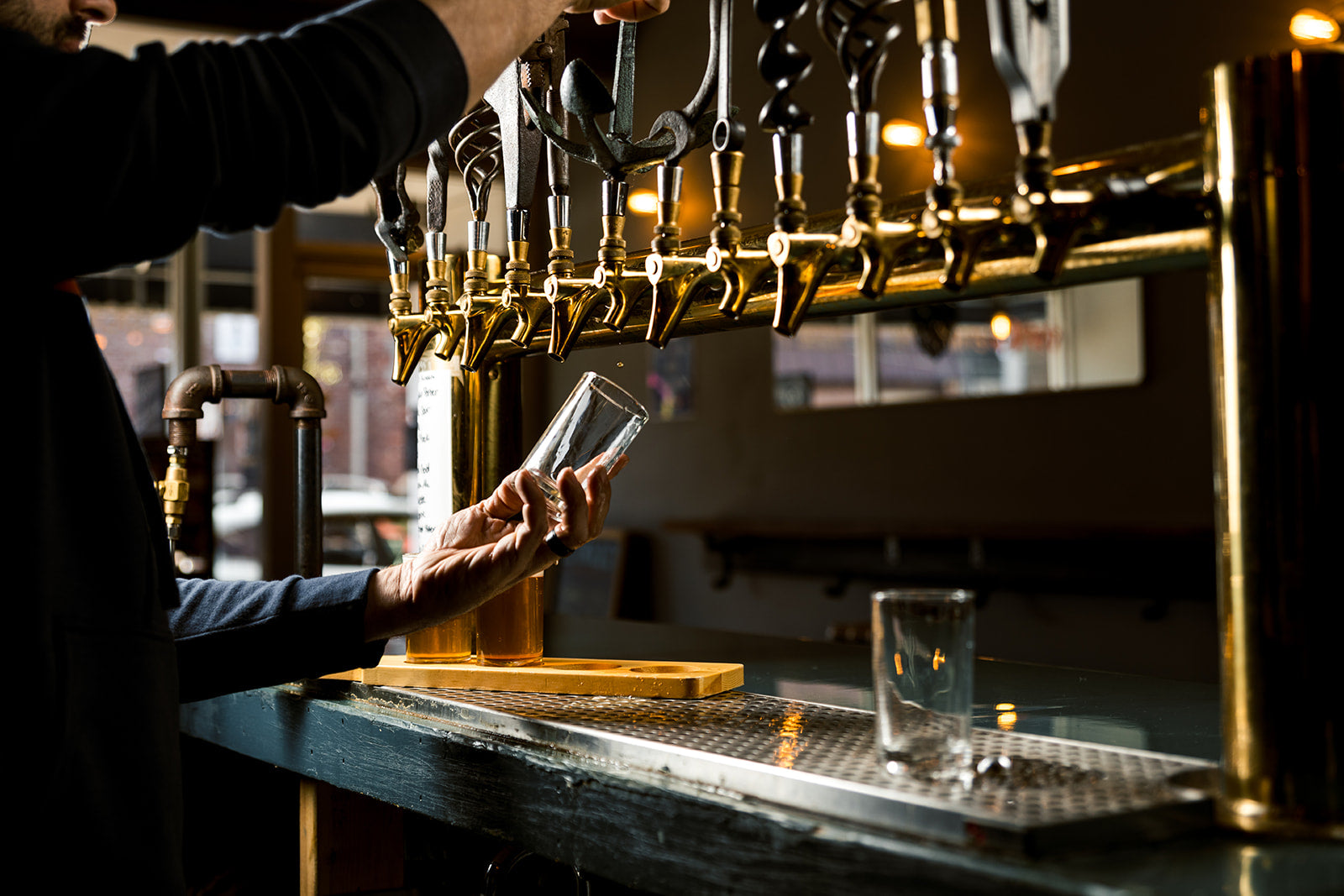 Load video: Man with flannel shirt walking through the tasting room padding the tap lines which beer is poured from. Man then stands in front of metal vessels and explains how beer is made.  Showing grain bags, beer cans, glasses of beers, also some clips of people filling the tasting room having some drinks and smiling